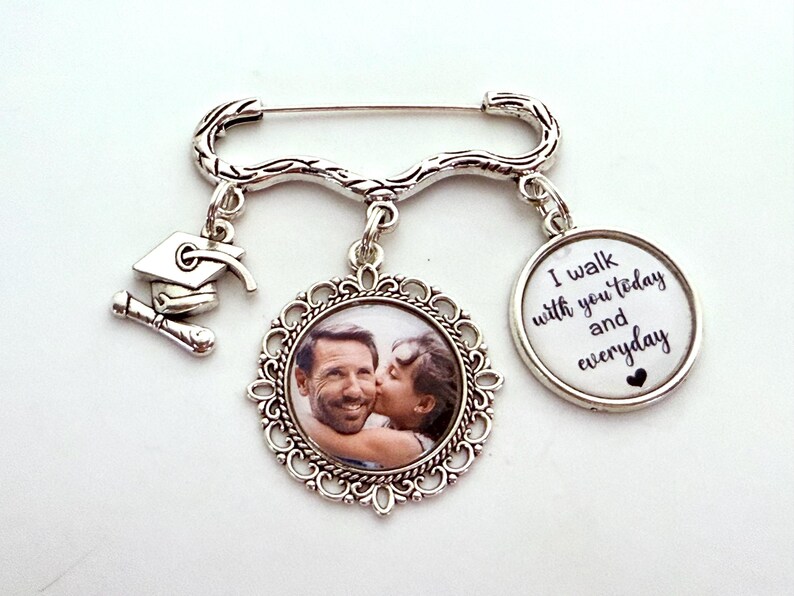 May include: Silver brooch with a graduation cap charm, a photo charm of a couple, and a charm with the text "I walk with you today and everyday".