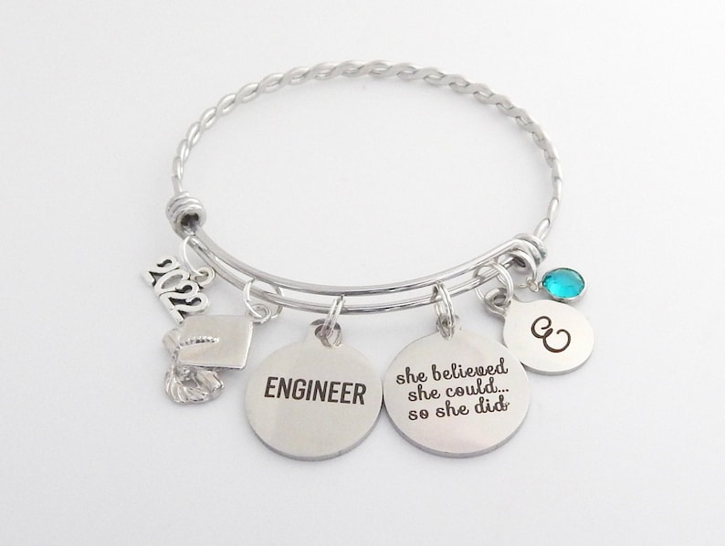 May include: Silver charm bracelet with a graduation cap charm, a charm that says "Engineer", a charm that says "She believed she could... so she did", a charm with the letter "C", and a turquoise blue crystal charm. The bracelet is braided and has a clasp.