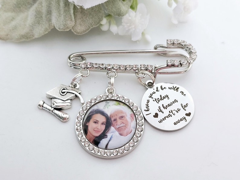 May include: Silver safety pin with a photo charm, a graduation cap charm, and a charm with the text "I know you'd be with me today if heaven weren't so far away".