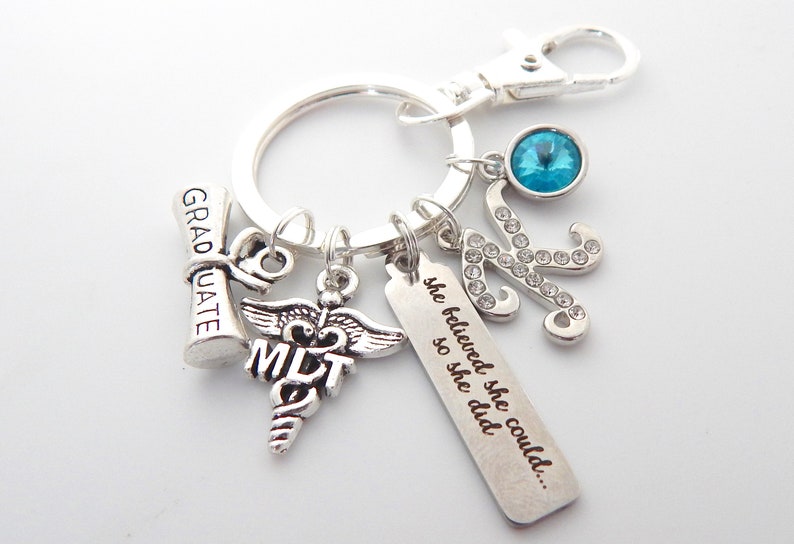 May include: Silver keychain with charms, including a graduation cap, a medical symbol with the letters MLT, a charm with the text "she believed she could... so she did", a rhinestone letter K, and a blue crystal charm.