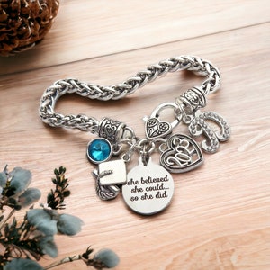 K&ouml;nnte beinhalten: Silbernes Armband mit einem Abschluss-Hut-Anh&auml;nger, einem Herz-Anh&auml;nger mit dem Wort "Klasse" darauf, einem blauen Kristall und einem Anh&auml;nger mit dem Text "Sie glaubte, sie k&ouml;nnte... also tat sie es".