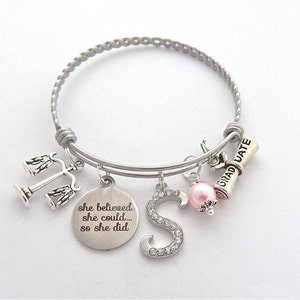 May include: Silver charm bracelet with a graduation cap, scales of justice, a pink pearl, and a charm that says "she believed she could... so she did".
