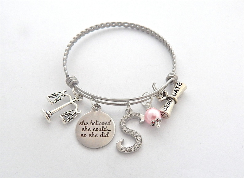 May include: Silver charm bracelet with a graduation cap, scales of justice, a pink pearl, and a charm that says "she believed she could... so she did".