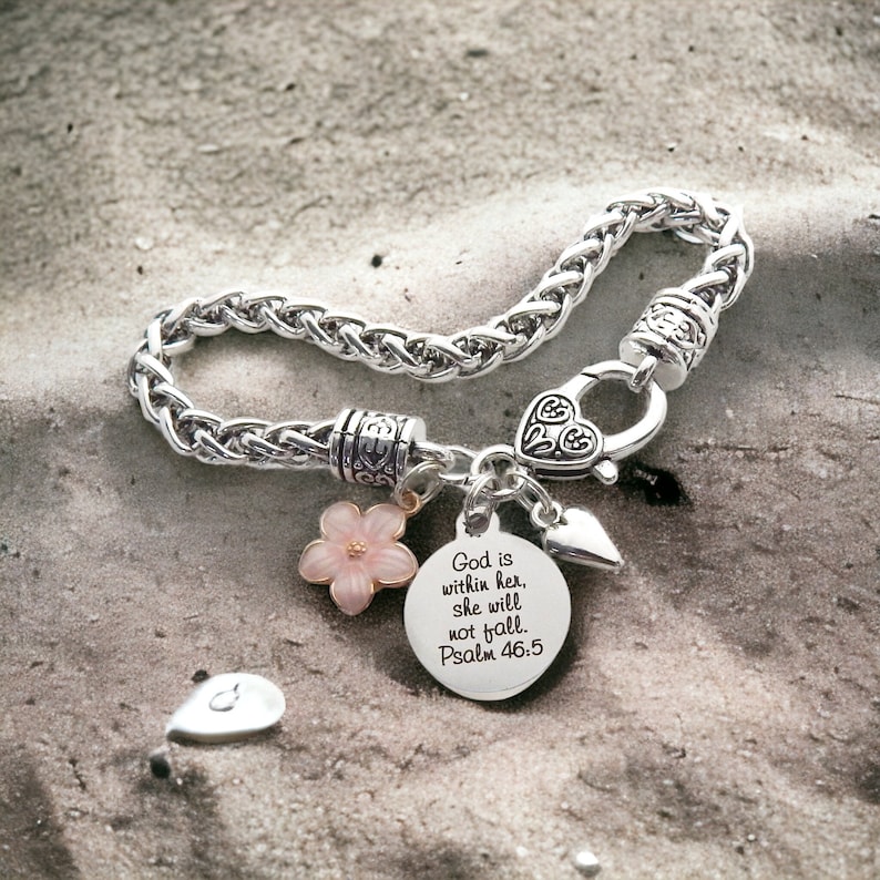 Puede incluir: Pulsera de plata con un dije que dice "Dios est&aacute; en ella, no caer&aacute;. Salmo 46:5". La pulsera tambi&eacute;n tiene un dije de flor rosa y un dije de coraz&oacute;n.