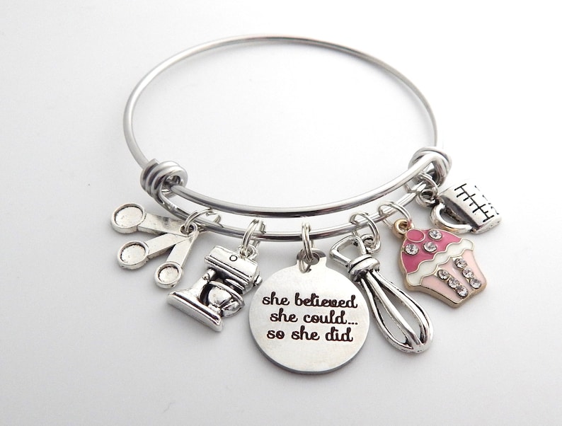 May include: Silver charm bracelet with a cupcake, measuring cup, whisk, measuring spoons, and a mixer charm. The charm with the text "She believed she could... so she did."
