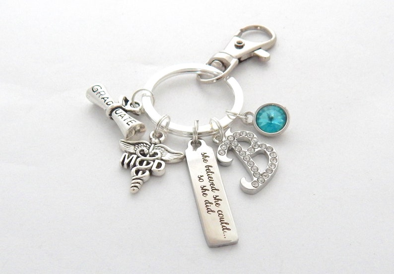 May include: Silver keychain with a graduation scroll charm, a medical symbol charm, a charm with the text "she believed she could... so she did", a rhinestone letter B charm, and a light blue rhinestone charm.