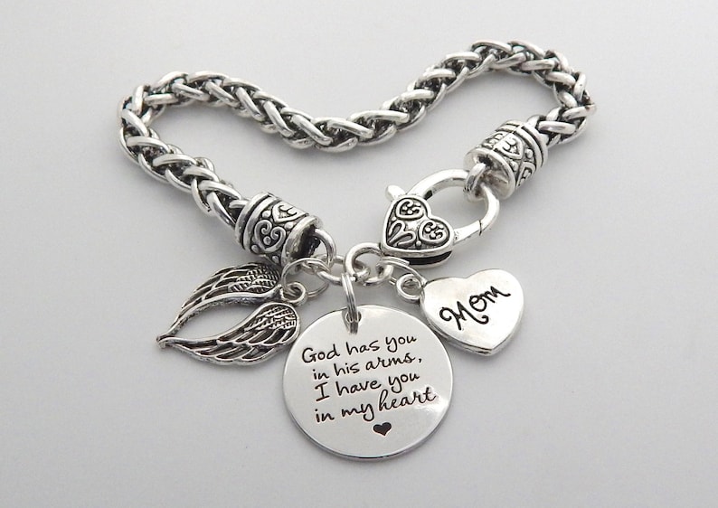 Op de afbeelding: Zilveren armband met een hartvormige bedel met de tekst "Mam" en een ronde bedel met de tekst "God heeft je in zijn armen, ik heb je in mijn hart". De armband heeft ook een paar engelenvleugels.
