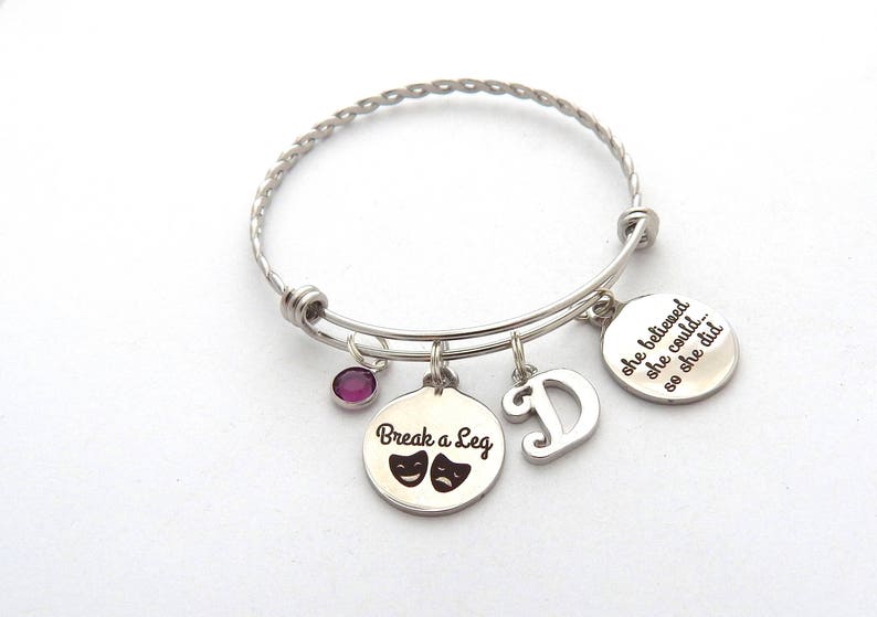 May include: Silver charm bracelet with a purple charm, a charm with the text "Break a Leg" and two theater masks, a charm with the letter "D", and a charm with the text "She believed she could... so she did."