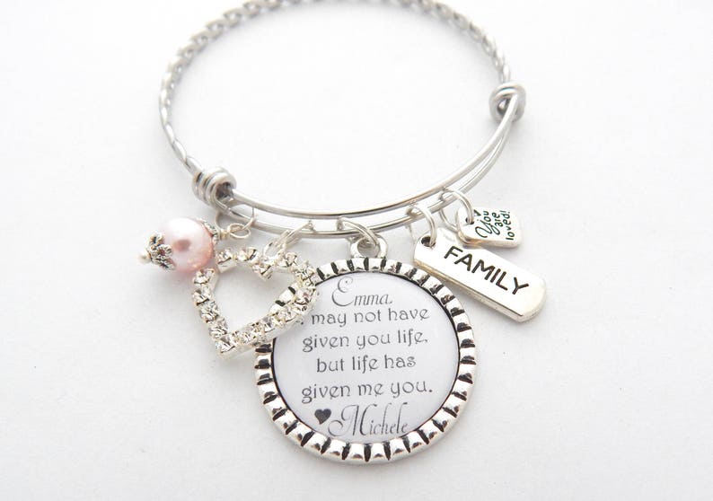May include: A silver charm bracelet with a heart charm, a pink pearl charm, and a round charm with the text "Emma. May not have given you life, but life has given me you. Michele" and a silver charm with the text "FAMILY".