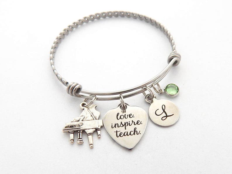 Peut inclure: Un bracelet &agrave; breloques en argent avec un charme de piano, un charme en forme de c&oelig;ur avec les mots "love, inspire, teach", un charme rond avec la lettre "L" et un charme en cristal vert.