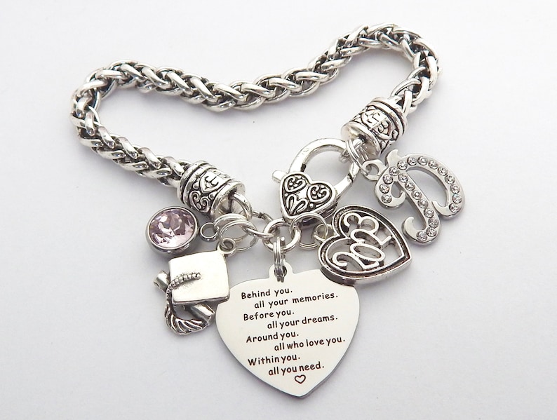 K&ouml;nnte beinhalten: Silbernes Charm-Armband mit einem Herz-Anh&auml;nger mit der Aufschrift "2023" und einem weiteren Herz-Anh&auml;nger mit einem inspirierenden Zitat. Das Armband hat au&szlig;erdem einen Abschluss-Hut-Anh&auml;nger, einen rosa Kristall-Anh&auml;nger und einen Buchstaben-Anh&auml;nger mit Strasssteinen.
