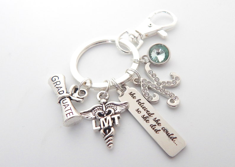 May include: Silver keychain with charms, including a graduation cap, a medical caduceus with the letters LMT, a charm with the text "she believed she could... so she did", and a silver letter K with rhinestones. The keychain also has a light blue crystal charm.