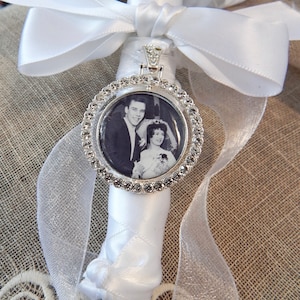 May include: A silver-toned circular photo charm featuring a black and white wedding portrait of a couple. The charm is bordered by clear rhinestones and is attached to a white satin ribbon wrapped around a stem, tied with a decorative bow.