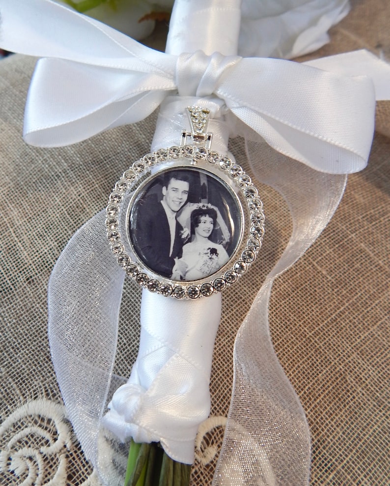 May include: A silver-toned circular photo charm featuring a black and white wedding portrait of a couple. The charm is bordered by clear rhinestones and is attached to a white satin ribbon wrapped around a stem, tied with a decorative bow.