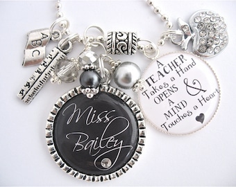 May include: A silver necklace with charms, including a ruler, a book with the letters ABC, a silver bead, a black bead, a silver bead, a silver charm with a swirl design, a white charm with the text "A Teacher Takes a Hand Opens a Mind & Touches a Heart", and a silver apple charm with rhinestones. The necklace also has a black charm with the text "Miss Bailey" in white.