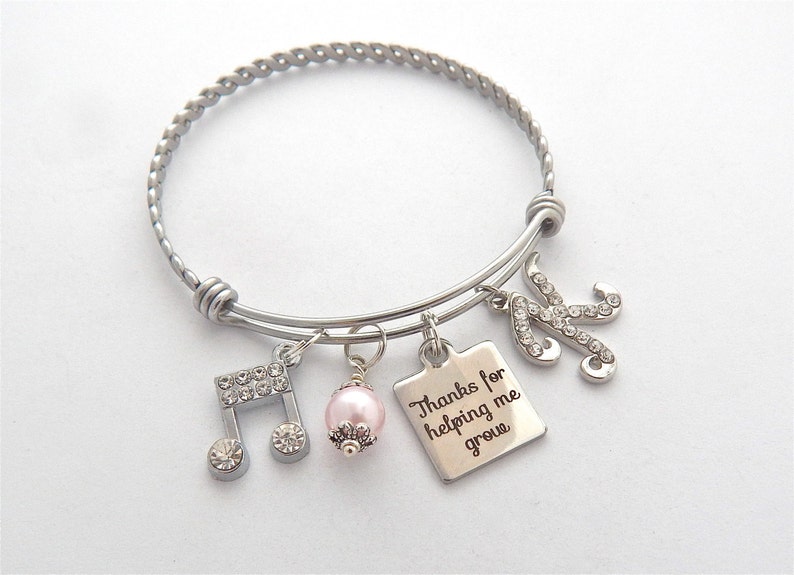 Peut inclure: Bracelet en argent avec un charme de perle rose, un charme de note de musique, un charme carr&eacute; avec le texte "Merci de m'aider &agrave; grandir" et un charme lettre K. Le bracelet est fait de fil d'argent torsad&eacute;.