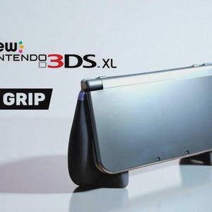 Könnte beinhalten: Eine silberne New Nintendo 3DS XL Konsole mit einem schwarzen Griffaufsatz. Die Konsole ist geschlossen und abgewinkelt, wobei die Ober- und Seitenansicht zu sehen sind. Der Text "new Nintendo 3DS XL" befindet sich über der Konsole, und das Wort "GRIP" steht in einem schwarzen Rechteck.