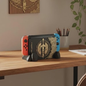 Puede incluir: Una consola Nintendo Switch negra con mandos naranja y azul, expuesta en un soporte negro. La consola presenta un diseño dorado en la parte trasera. El soporte está sobre una superficie de madera.