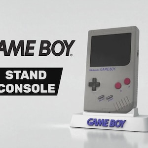 Peut inclure: Une console Game Boy grise posée sur un support blanc avec le mot "GAME BOY" en bleu. La console a un écran noir, une croix directionnelle, des boutons roses et le texte "Nintendo GAME BOY". Le fond affiche "GAME BOY STAND CONSOLE".