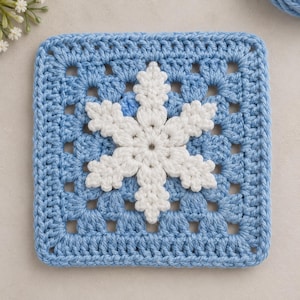 May include: A square crochet granny square featuring a light blue yarn base with an intricate, raised white snowflake motif centered on the surface. The design displays a classic open-work stitch pattern with a textured, hand-crafted appearance.