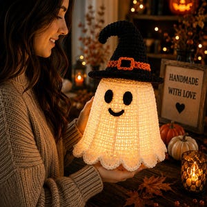 May include: A crocheted ghost figurine featuring a white body with black eyes and a black mouth, wearing a black witch hat with an orange band and buckle. The ghost is illuminated from within, casting a warm glow through the textured yarn stitches.