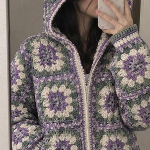 May include: A zip-up hooded jacket constructed from crocheted granny squares in a repeating pattern of lavender, sage green, and cream yarn. The garment features a full-length front zipper, long sleeves, and a hood, showcasing a textured, handmade aesthetic.
