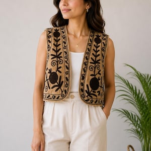 Puede incluir: Un chaleco sin mangas de color tostado con patrones florales y de enredaderas bordados en marrón oscuro en los paneles frontales. El chaleco tiene un escote redondeado y un diseño abierto, usado sobre una camiseta blanca y pantalones claros.