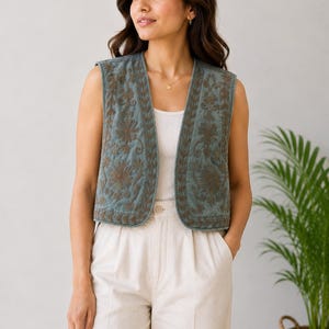 Può includere: Un gilet senza maniche in una tonalità blu-verde spenta con intricati ricami floreali marroni sui pannelli frontali. Il capo presenta un'apertura frontale dritta, uno scollo arrotondato e una consistenza trapuntata, indossato sopra una canotta bianca.