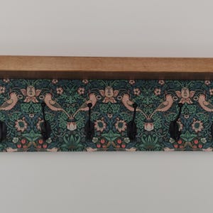 Può includere: Una mensola da parete in legno con sei ganci neri. La mensola ha un piano in legno naturale e lati dipinti di verde acqua. Lo schienale presenta un motivo floreale con uccelli nei toni del rosa, verde e verde acqua.