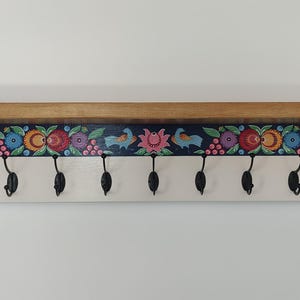 May include: Wooden wall shelf with a light brown finish, featuring a decorative floral and bird design in vibrant colors. The shelf includes eight black metal hooks for hanging items. The design is painted on a dark blue background.