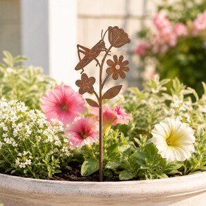 Pode incluir: Uma estaca de jardim de metal cor de ferrugem com a silhueta de um gafanhoto pousado num caule com três flores estilizadas e duas folhas, inserida num vaso redondo cheio de flores cor-de-rosa, brancas, amarelas e folhagem verde.