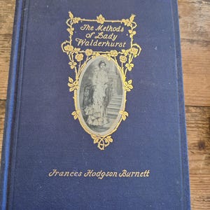 Puede incluir: Un libro antiguo de tapa dura titulado "The Methods of Lady Walderhurst" de Frances Hodgson Burnett. El libro tiene una cubierta azul oscuro con letras doradas y un marco dorado adornado alrededor de un retrato de una mujer.