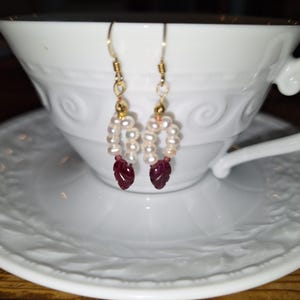 May include: A pair of dangle earrings featuring a gold-toned hook, a small gold bead, a circular arrangement of white freshwater pearls, and a single deep red, carved leaf-shaped gemstone pendant hanging at the base of each earring.