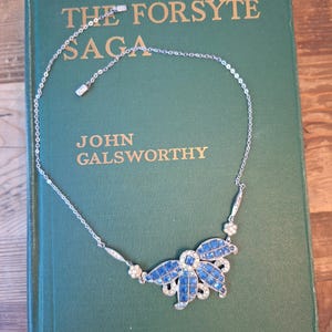 May include: A silver-toned necklace featuring a central floral-shaped pendant adorned with square-cut blue stones and small clear crystals. The delicate chain includes small decorative metal links and a secure clasp, resting on a green book cover.