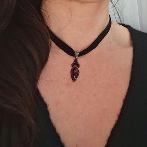 May include: A black velvet choker necklace featuring a teardrop-shaped dark red gemstone pendant. The pendant is framed by a cluster of smaller, matching red stones and is attached to the band with a simple silver-toned metal bail.
