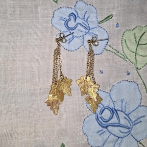 May include: A pair of gold-toned dangle earrings featuring multiple delicate chains suspended from a stud post. Each earring terminates in a cluster of three textured, leaf-shaped charms that hang at varying lengths, creating a layered effect.