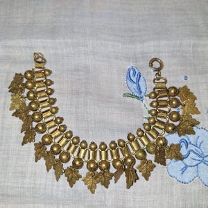 May include: A gold-toned metal charm bracelet featuring a series of repeating spherical beads and flat, textured leaf-shaped charms dangling from a central chain link structure, secured with a circular spring ring clasp at one end.