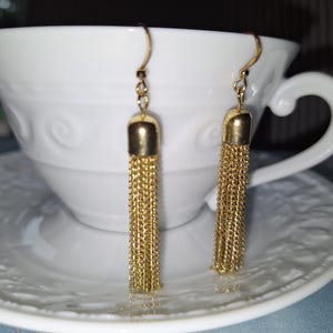 May include: A pair of gold-toned dangle earrings featuring a polished metal cap at the top and a long, multi-strand chain tassel hanging below. Each earring is attached to a simple curved ear wire hook for secure placement in the earlobe.