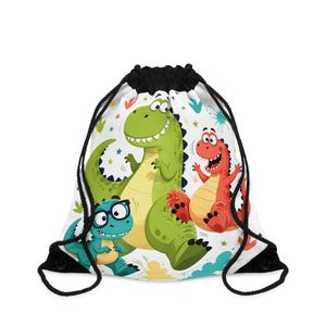 Puede incluir: Una mochila de cuerdas blanca con un estampado de dibujos animados de tres dinosaurios. Un gran dinosaurio verde se encuentra en el centro, un dinosaurio rojo más pequeño está a la derecha y un pequeño dinosaurio azul verdoso con gafas a la izquierda.