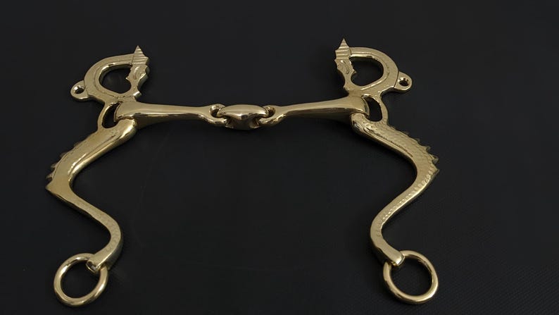 May include: A gold-coloured horse bit with a curved design. The bit has a central joint and rounded rings on each end. The design includes decorative elements near the top and along the curved sides. The background is black.