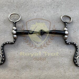 May include: A horse bit with a black and brown center bar and silver-colored rings. The bit has a curved design with small silver studs along the sides. The bit is designed for equestrian use.