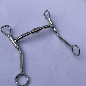 May include: A stainless steel horse bit with a copper centre piece. The bit has a curved design with rings on each end and a jointed mouthpiece. The bit is designed for use in equestrian activities.