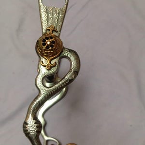 Puede incluir: Un bocado de caballo plateado y dorado con intrincados diseños florales y de hojas. El bocado presenta una forma curva y serpentina con una pieza central dorada decorativa. El bocado está diseñado para su uso en actividades ecuestres.