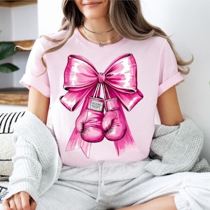 May include: A light pink short-sleeve t-shirt featuring a large, vibrant pink bow graphic tied around a pair of pink boxing gloves. The boxing gloves have a small white label on the wrist area with the text "BOXING HOPE" printed in black.