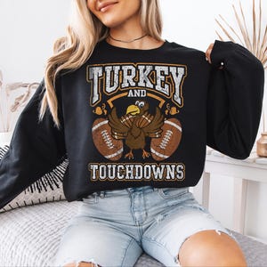 May include: A black long-sleeve sweatshirt featuring a graphic print with the text TURKEY AND TOUCHDOWNS in a distressed white font. The design includes a central cartoon turkey with brown feathers and two brown footballs on either side.