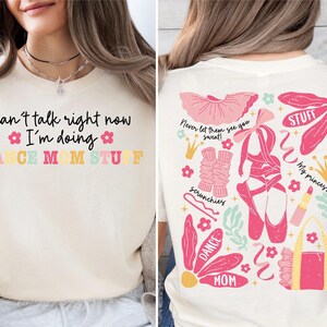 May include: A cream-colored t-shirt featuring black text on the front that reads Can't talk right now I'm doing DANCE MOM STUFF. The back displays a colorful graphic print of ballet shoes, a tutu, leg warmers, a lipstick, and the text Never let them see you sweat.