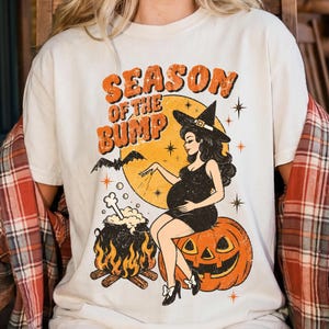 Może przedstawiać: Kremowy t-shirt z nadrukiem graficznym z napisem "SEASON OF THE BUMP" w kolorze pomarańczowym. Wzór przedstawia ciężarną czarownicę siedzącą na dużej dyni, bulgoczący kociołek z kością, żółty księżyc oraz nietoperza.