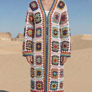 May include: A long, open-front crochet cardigan in a patchwork design. The cardigan features a white base with colorful squares in shades of blue, red, yellow, and brown. The edges are trimmed with blue and rainbow-colored yarn, creating a bohemian aesthetic.