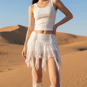 May include: A white tank top and a white lace skirt with asymmetrical hemline. The skirt has a layered design. The person is wearing cream-colored boots. The background is a desert landscape with sand dunes and a clear blue sky.