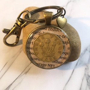 May include: A bronze-toned keychain with a circular pendant displaying a 1939 Three Pence coin. The pendant is set in a bronze frame, with a small heart charm. The background features text from a book.